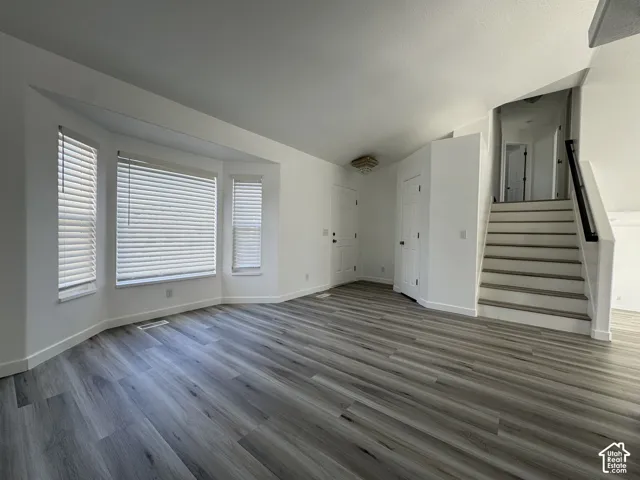 Bay window with blinds, wood-finish flooring, staircase with dark railing, white walls, and a ceiling light fixture
