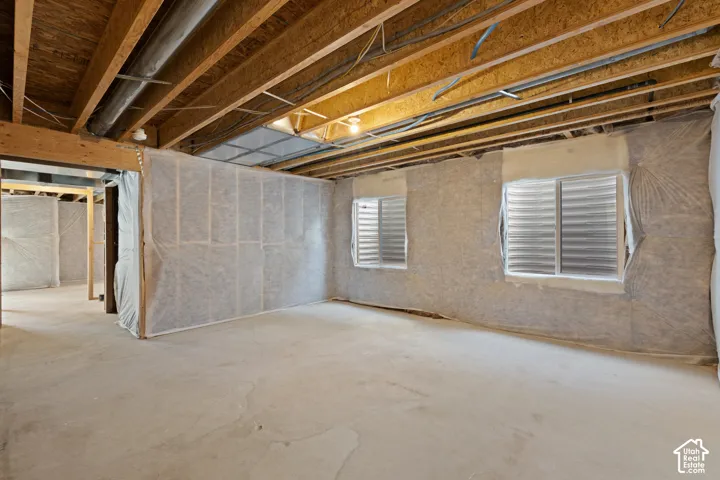 Exposed wood ceiling joists with visible electrical and plumbing infrastructure, concrete flooring, and two small windows with louvered blinds
