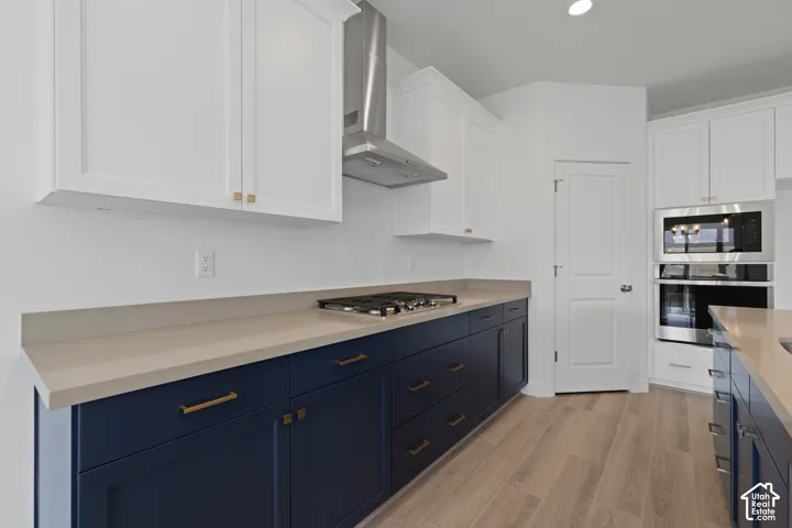 Kitchen featuring two-tone cabinetry, light-toned countertops, a stainless steel range hood, built-in microwave and oven, and wood-finish flooring