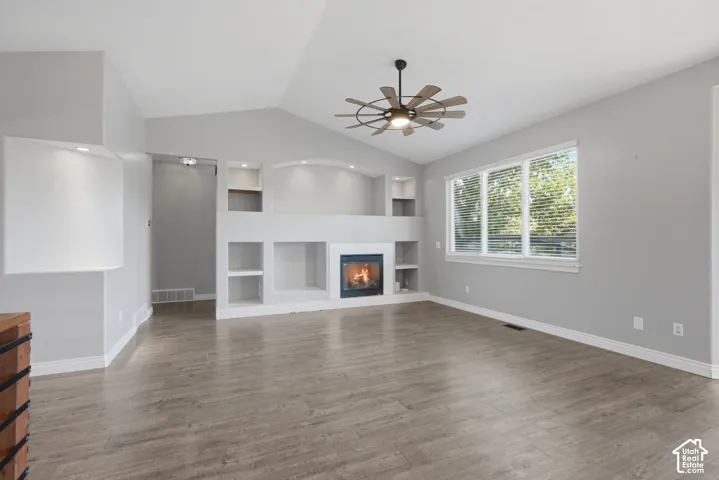 Vaulted ceiling with an overhead fan, built-in fireplace, recessed shelving, large window with blinds, and wood-finish flooring