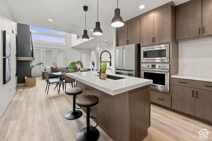Modern kitchen featuring custom wood-finish cabinetry, white quartz waterfall island with undermount sink, stainless steel appliances including dual wall ovens and microwave, pendant lighting, and light wood-finish flooring extending into the open dining