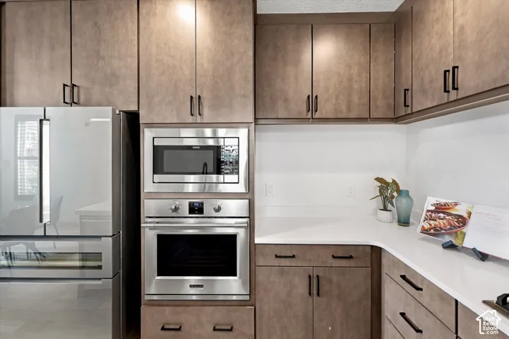 Modern kitchen featuring stainless steel French-door refrigerator, built-in microwave and wall oven, white quartz countertops, and wood-finish cabinetry with black hardware throughout