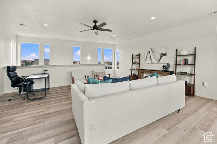 Bright, open living area featuring expansive windows with natural light, wood-finish flooring throughout, a ceiling fan with remote control, built-in shelving with ladder accent, and a spacious sectional layout ideal for entertaining