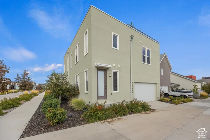 Modern two-story residence featuring a clean-lined sage exterior with large window openings, an attached two-car garage, concrete driveway and walkway, and landscaped front grounds with shrubs and ground-level plantings