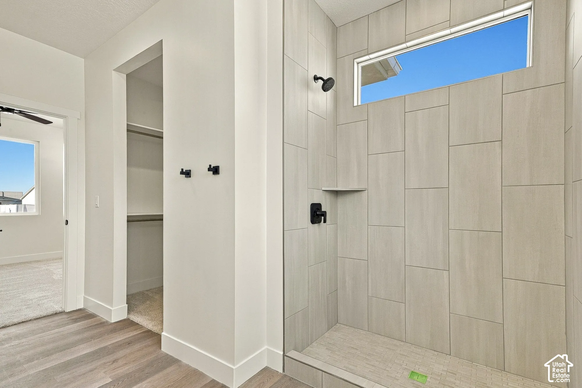 Walk-in shower featuring large-format neutral wall tile, integrated corner shelving, matte black fixtures, and a horizontal transom window