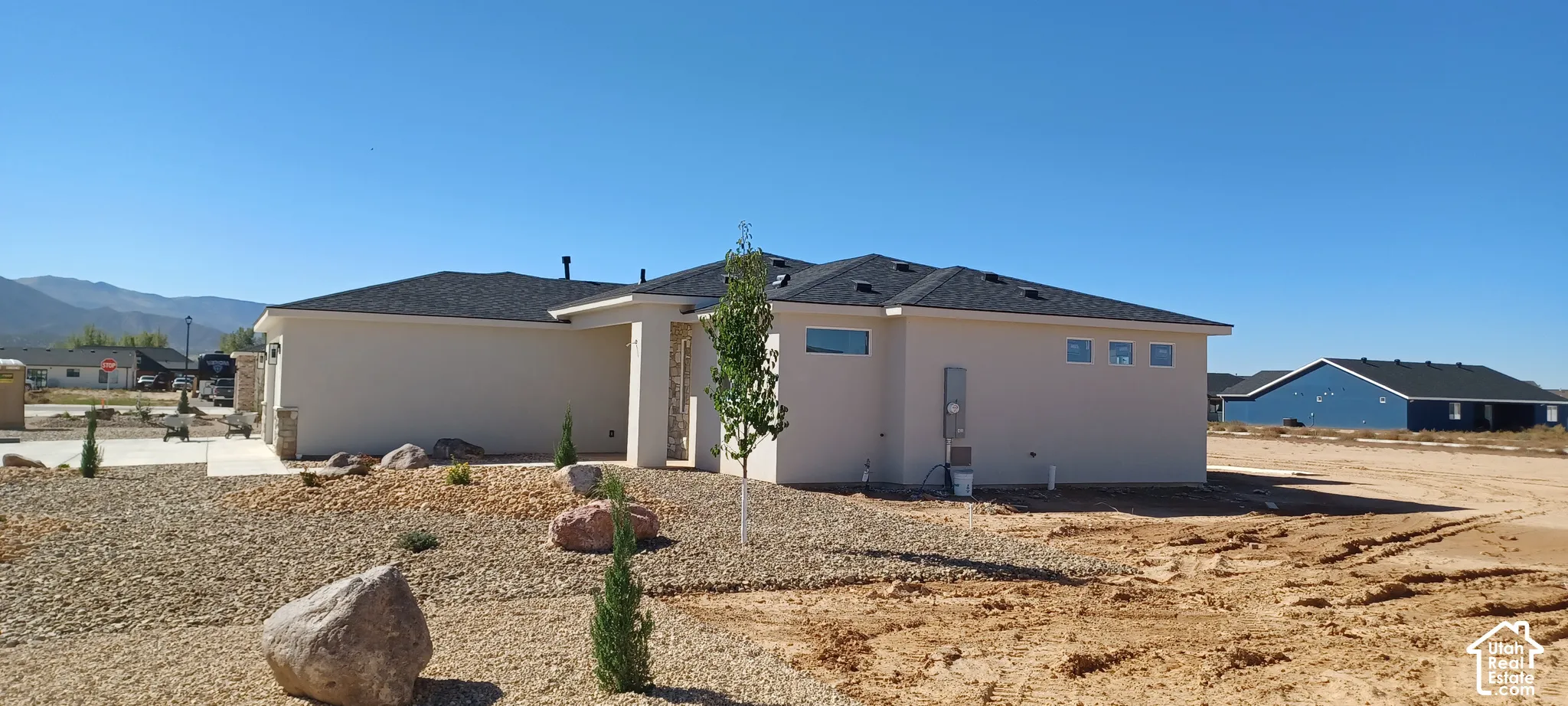 Single-story residence featuring a smooth stucco exterior, dark shingle roof, stone pillar accents, and rock landscaping