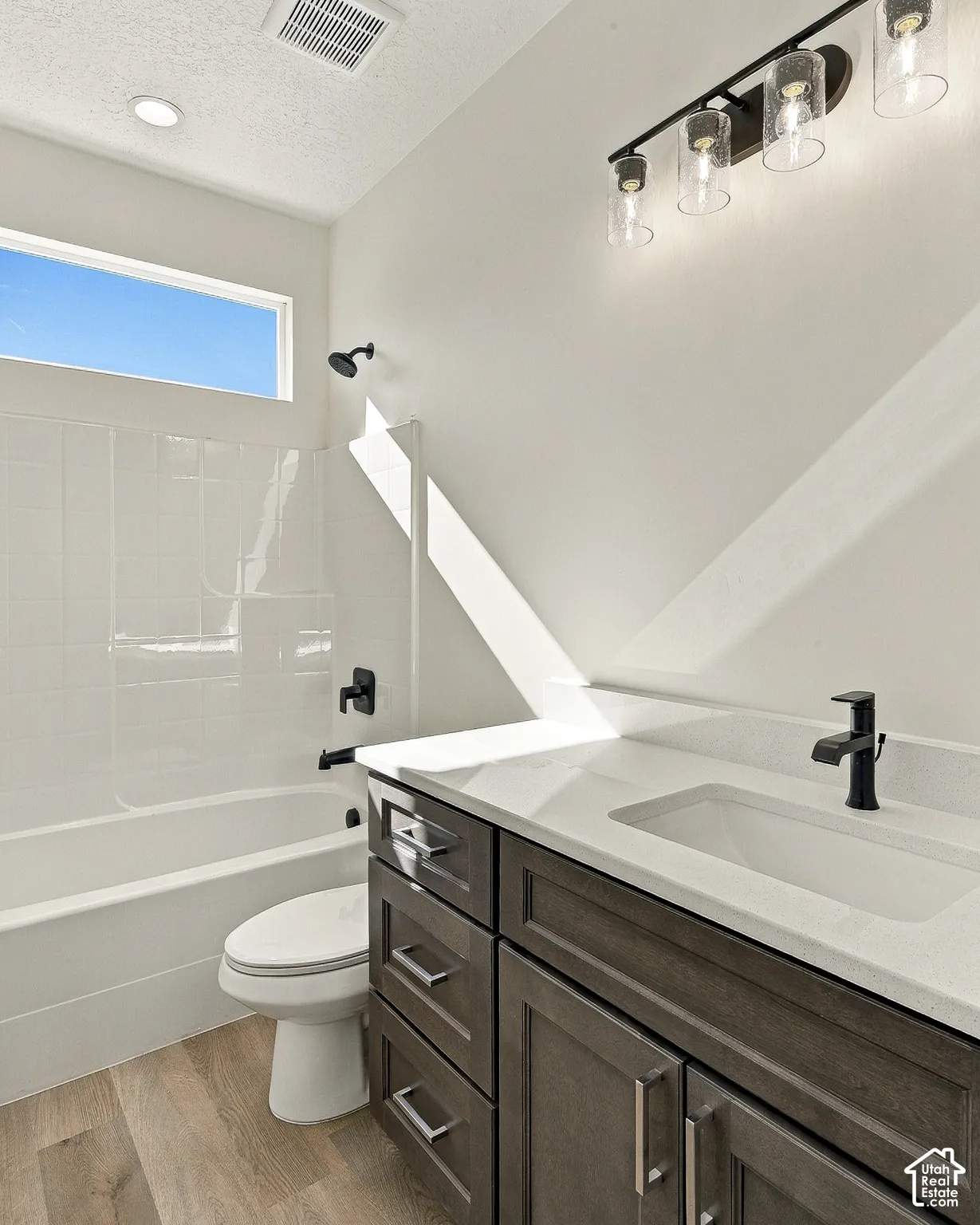Bathroom featuring a vanity with dark-stained cabinetry, a light-toned countertop, and matte black hardware, paired with wood-finish flooring, a porcelain tub surround, and a four-light glass fixture