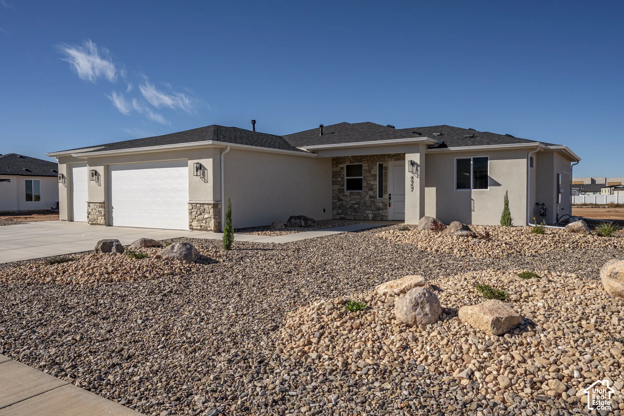 Single-story exterior featuring a neutral stucco finish, stacked stone accents, shingle roof, two-car garage, and xeriscaped lot with decorative boulders