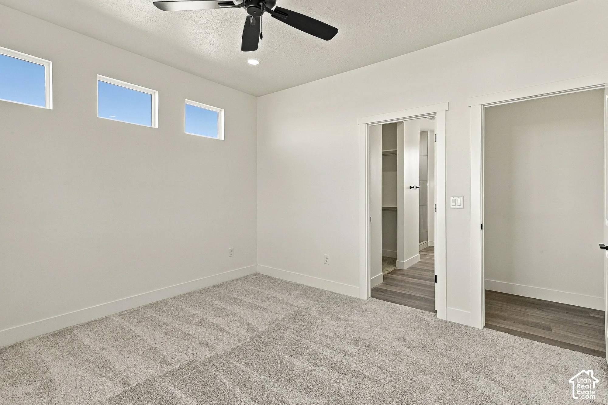 Room featuring neutral carpeting, three high-set transom windows, a matte black ceiling fan, and access to walk-in storage with wood-finish flooring