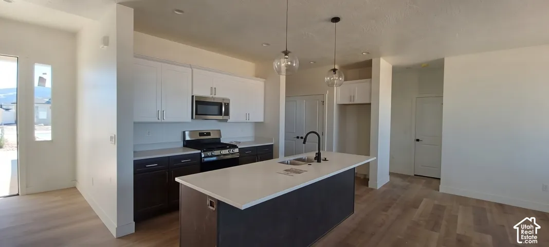 Kitchen featuring an island with sink, stainless steel appliances, upper white cabinetry, lower dark cabinetry, and wood-finish flooring