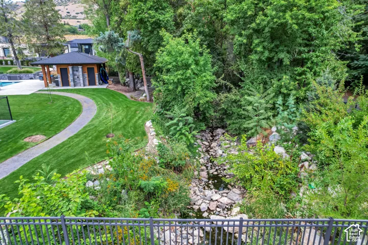 Manicured lawn with a winding pathway, adjacent to a wooded area featuring a natural stream, and a built-in pool house with stone accents