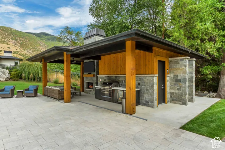 Expansive paver patio with a covered outdoor kitchen, built-in fireplace, natural stone accents, and wood paneling