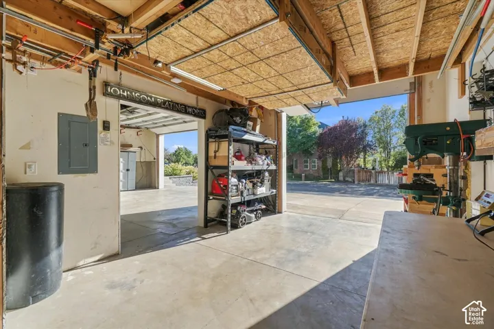 Concrete flooring, exposed wood beam ceiling, overhead fluorescent lighting, open garage door, and visible electrical panel