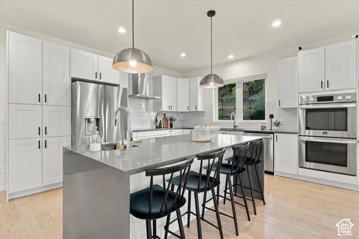 Kitchen featuring light wood-finish flooring, white cabinetry, a central island with a waterfall countertop, stainless steel appliances, and pendant lighting