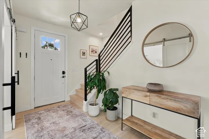 Light wood-finish flooring, white paneled entry door with transom windows, black geometric pendant light, and black metal stair railing