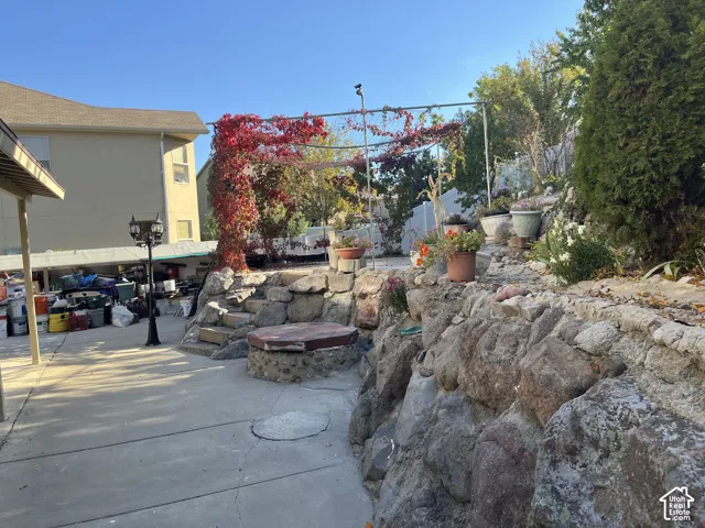 Paved patio area, stone retaining wall, tiered landscaping, and an overhead trellis with red foliage