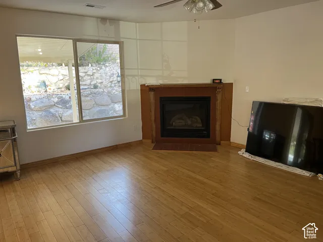 Engineered wood flooring, corner fireplace with wood surround and mantel, built-in ceiling fan, and large windows with natural light