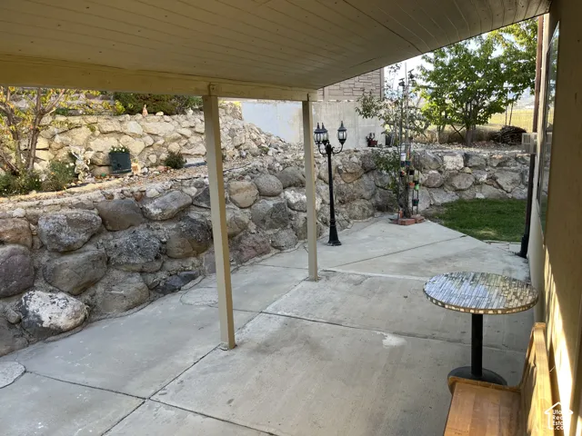 Covered patio with concrete flooring, supported by structural posts, and featuring a stone retaining wall