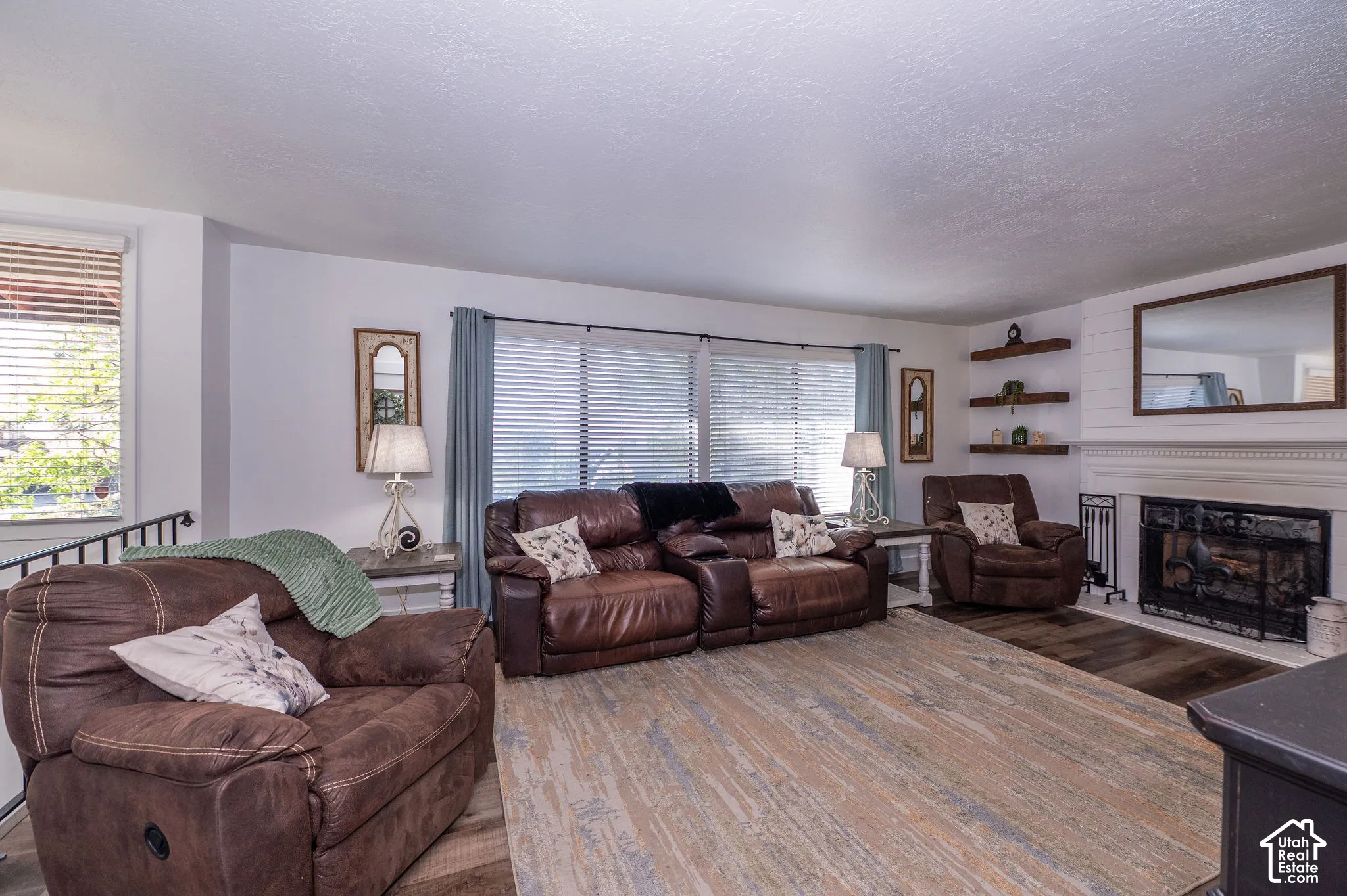 Spacious living area featuring large windows, a fireplace with a white mantel, floating shelves, and wood-finish flooring