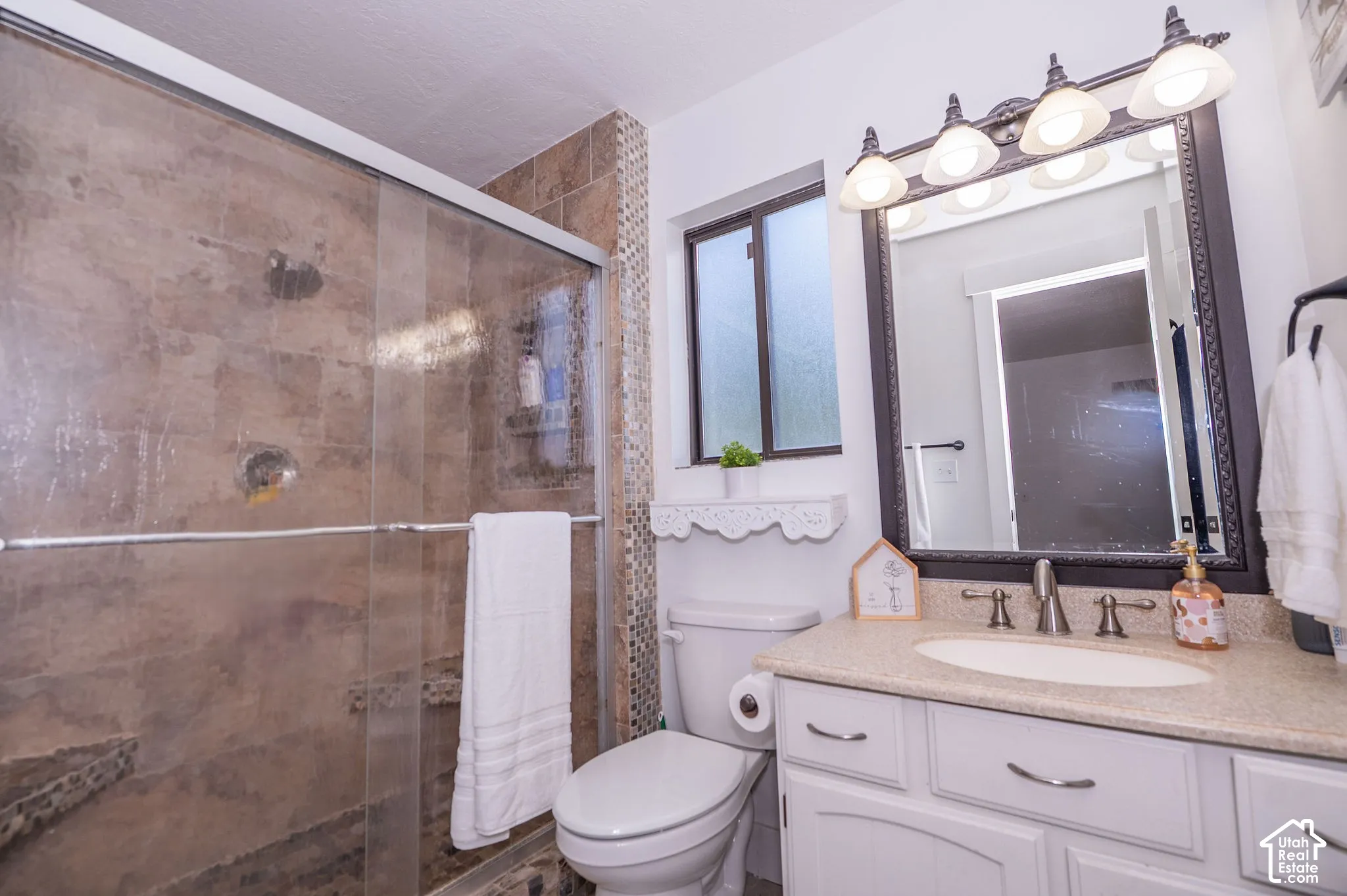 Primary ensuite tiled shower enclosure with sliding glass door, white vanity with integrated sink, rectangular framed mirror with sconce lighting, and a frosted window