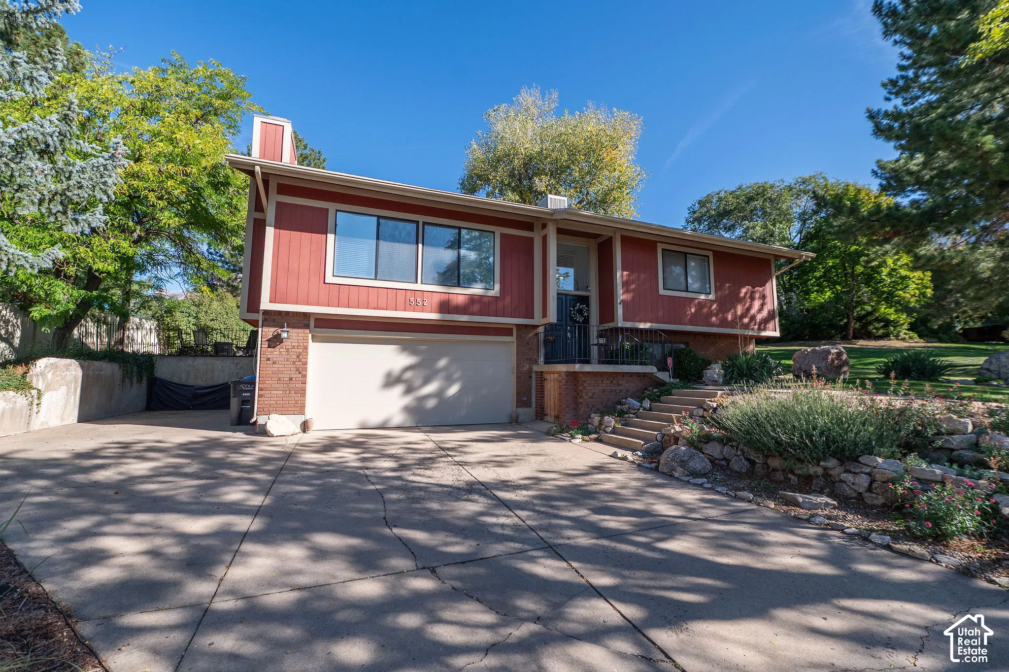 Red exterior siding with brick accents, attached garage, concrete driveway, stone retaining walls, and mature landscaping