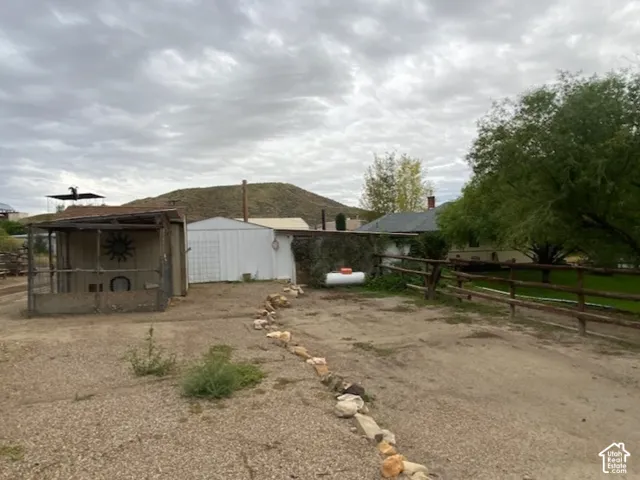 Back side of the out building showing the propane tank for the fireplace. Gravel driveway with a rock-lined border, wooden fence, and mature trees