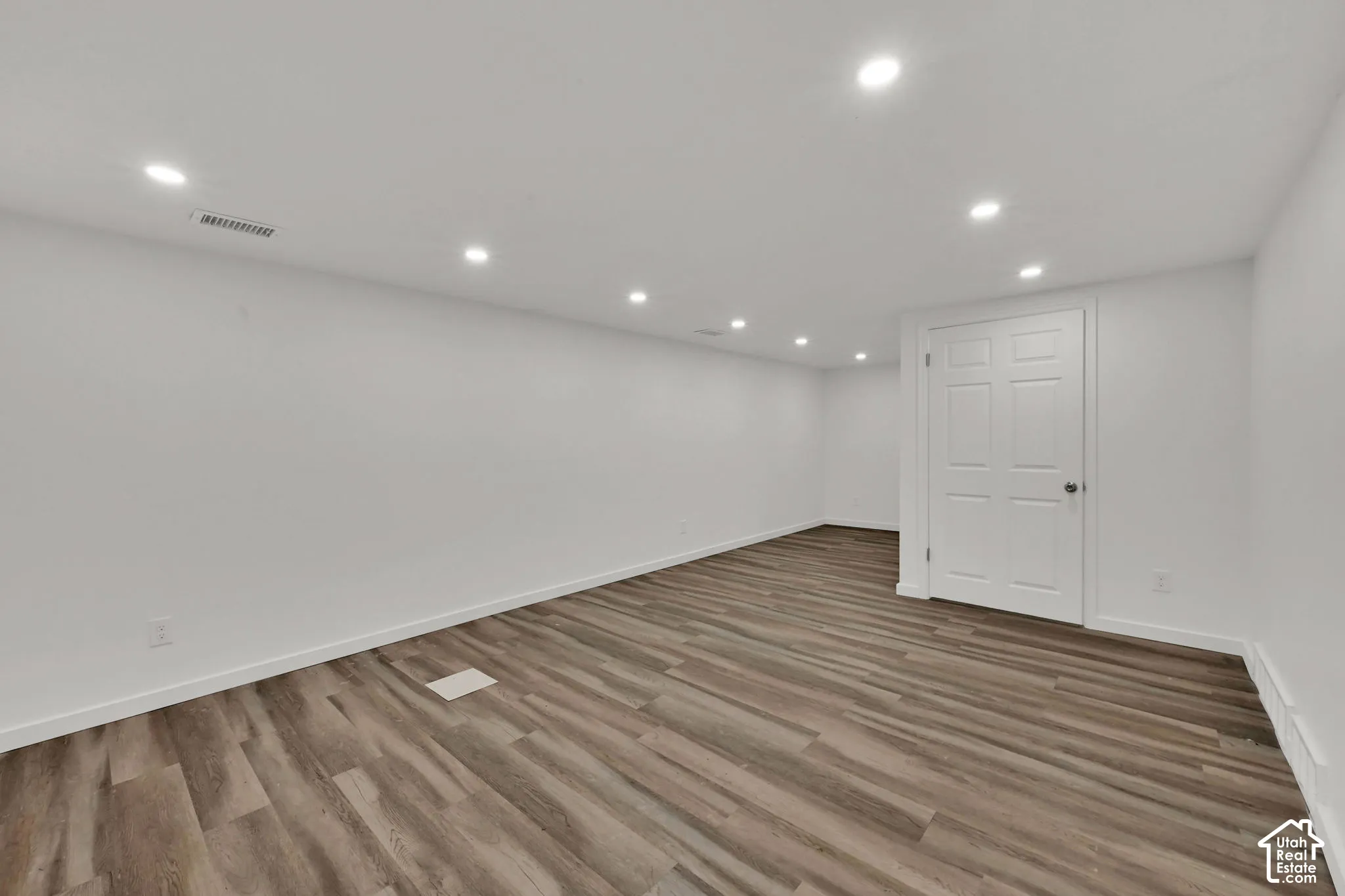 Spacious room with recessed lighting, wood-finish flooring, white walls, and a paneled door