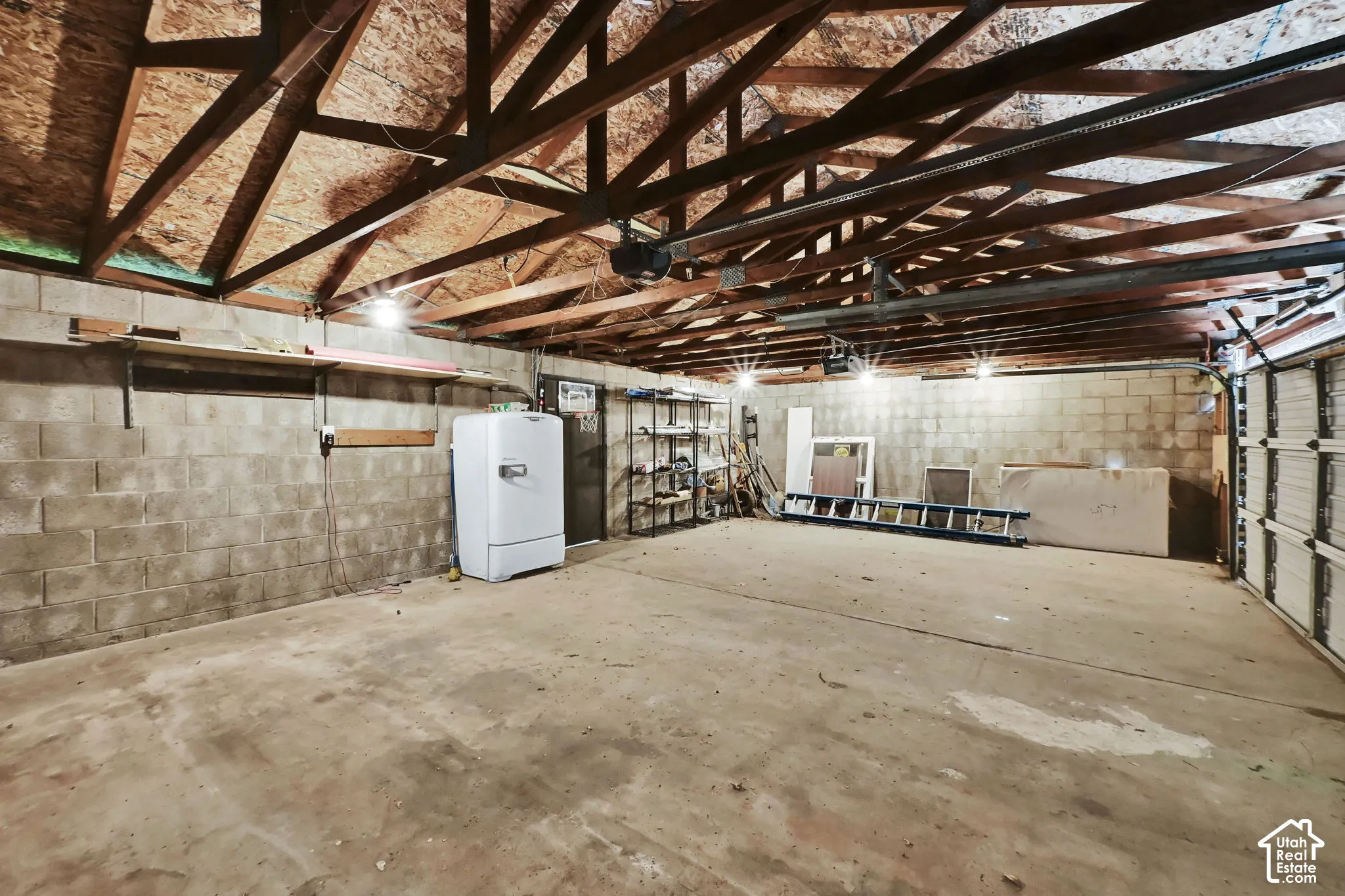 Exposed rafter ceiling, concrete block walls, concrete flooring, overhead lighting, and an automated garage door