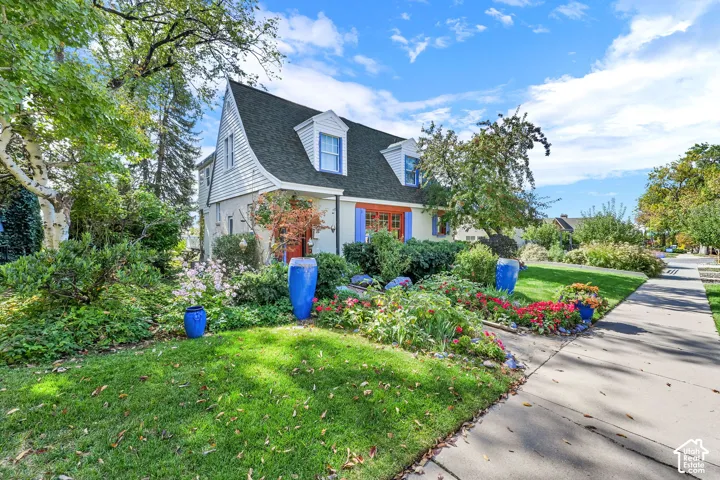 Gable roofline with dormer windows, surrounded by mature trees, and a landscaped front yard featuring diverse plantings and vibrant flowers