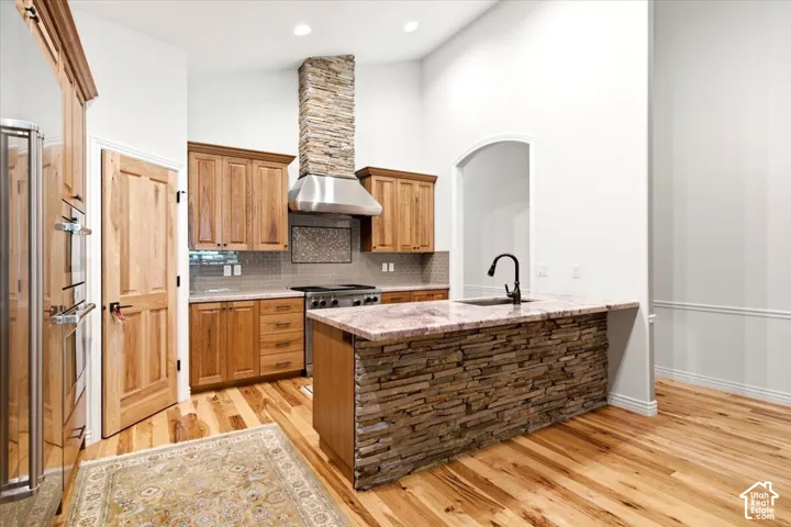 Kitchen featuring a stone-clad island with a sink, wood cabinetry, stainless steel range hood, and wood flooring