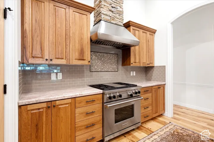 Stainless steel range with a ventilation hood, stacked stone range surround, wood cabinetry, light-toned countertops, and subway tile backsplash