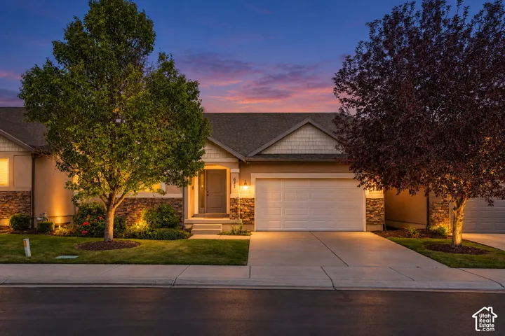Warm exterior lighting, stone accent facade, attached garage, concrete driveway, and a well-maintained lawn