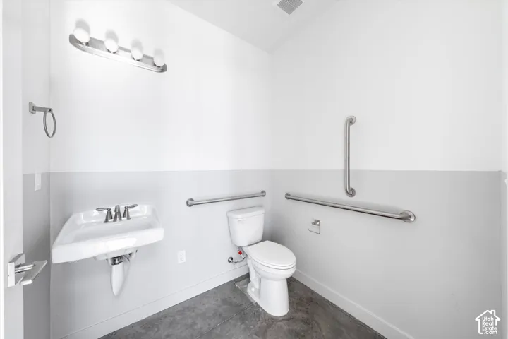 White wall-mounted sink with chrome-finish faucet, toilet with exposed plumbing, concrete-finish flooring, and multiple grab bars