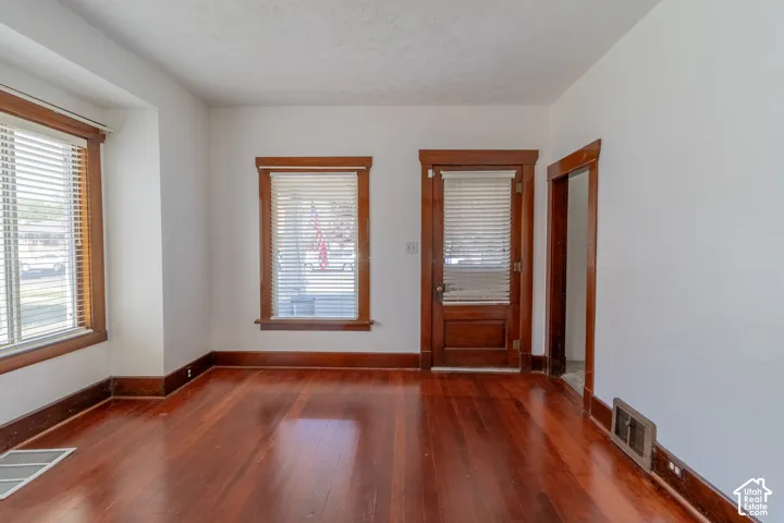 Hardwood-finish flooring, white painted walls, wood-trimmed windows with blinds, a wood door, and a doorway
