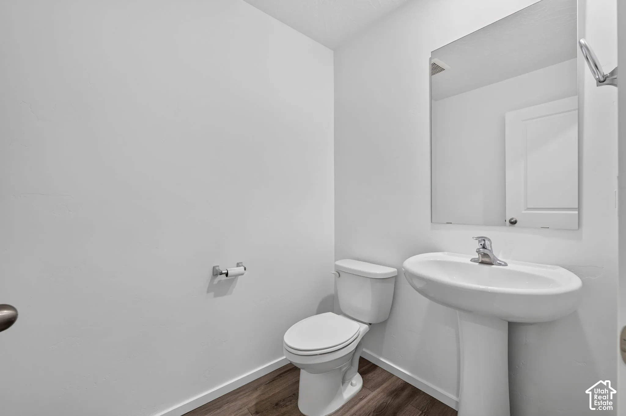 Pedestal sink with chrome faucet, wall-mounted mirror, white toilet, and wood-finish flooring