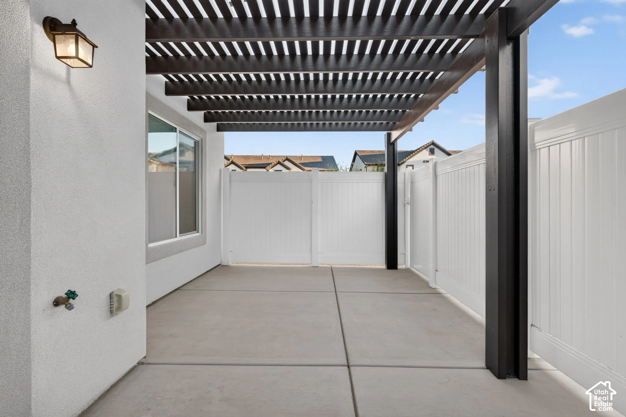 Patio area featuring a slatted pergola, concrete flooring, exterior wall lantern, and white privacy fencing