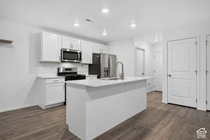 Kitchen featuring white cabinetry, stainless steel appliances, a center island with sink, white countertops, and wood-finish flooring