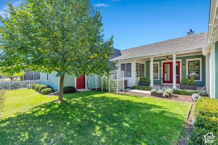 Covered porch with column supports, red entry door, exterior with a light blue finish, front yard with mature landscaping, and a white picket fence