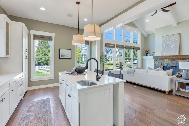 Vaulted ceilings with exposed beams, stone fireplace with wood mantel, large windows, wood-finish flooring, and a kitchen island with integrated sink