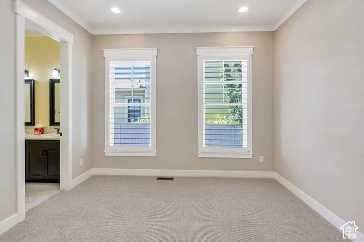 Recessed lighting, crown molding, two windows with plantation shutters, light-colored carpet flooring, and an adjoining bathroom with a dark wood vanity and dual mirrors