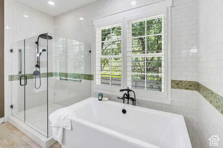 Soaking tub with wall-mounted faucet, walk-in shower with glass enclosure, white subway tile surround, accent tile detailing, and plantation shutters
