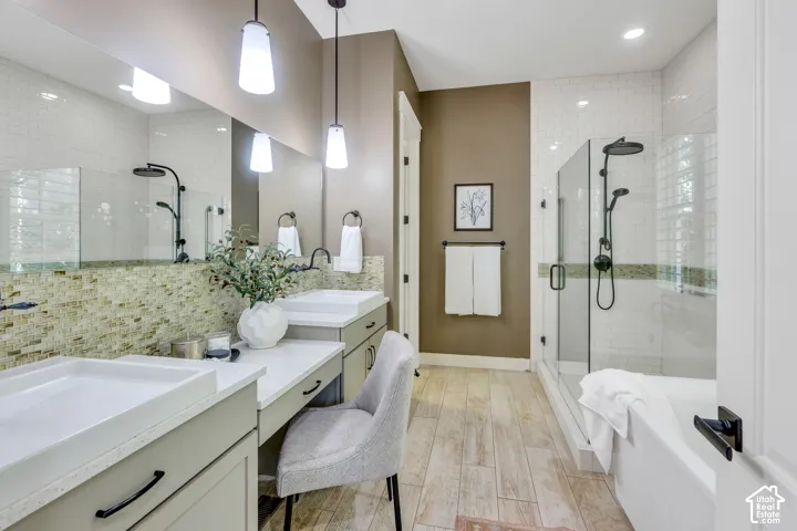 Dual vanity with integrated sinks, mosaic tile backsplash, wood-finish flooring, frameless glass shower enclosure, and a freestanding soaking tub