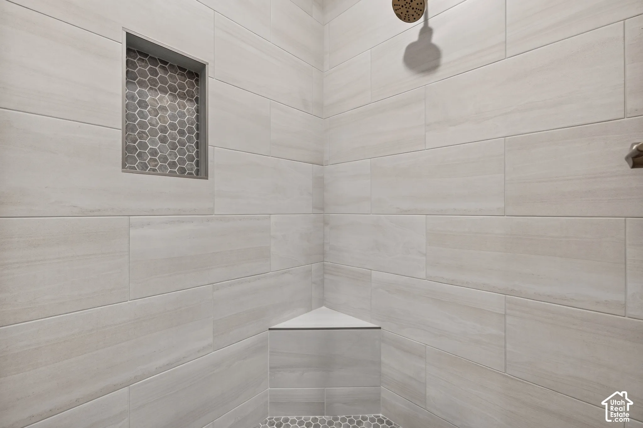 Shower featuring light-toned rectangular wall tiling, a built-in corner bench, a recessed niche with honeycomb mosaic tiling, and a bronze-finish showerhead
