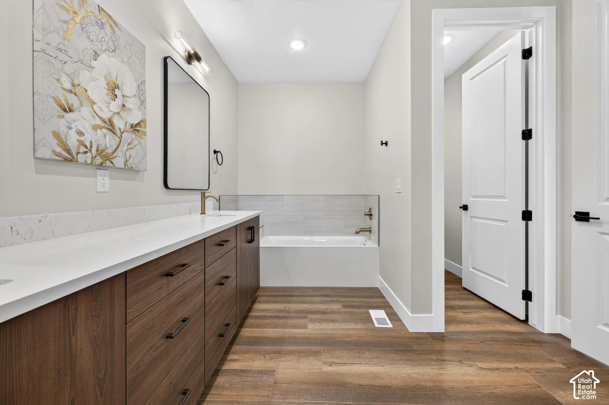 Wood-finish flooring, a double vanity with stone countertops and dark wood cabinetry, a recessed bathtub with tile surround, and a rectangular mirror with sconce lighting