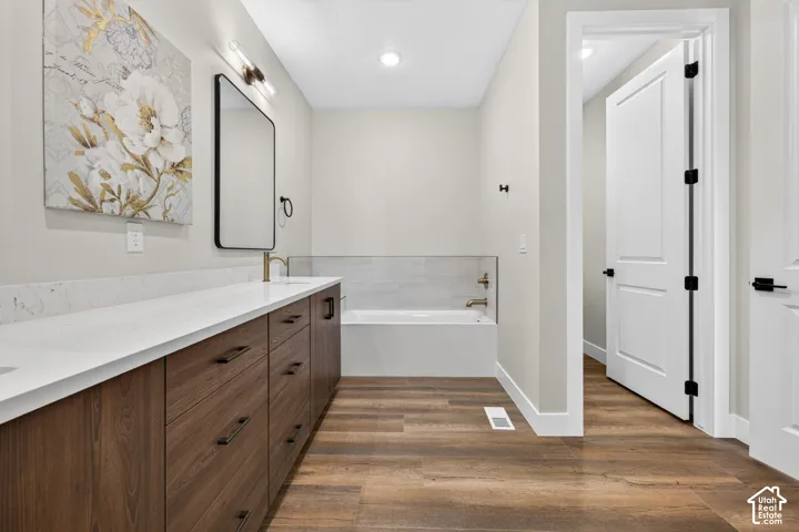 Wood-finish flooring, a double vanity with stone countertops and dark wood cabinetry, a recessed bathtub with tile surround, and a rectangular mirror with sconce lighting