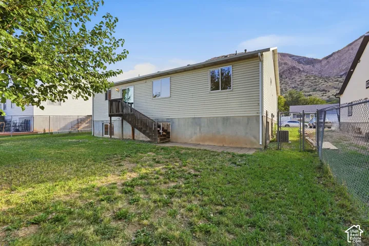 Rear exterior featuring light-colored siding, multiple windows, a wooden deck with stairs, a chain-link fence, and a distant mountain backdrop