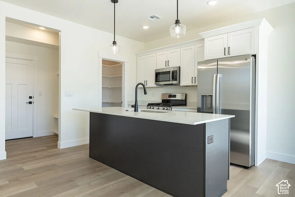Modern kitchen featuring white shaker cabinetry, stainless steel appliances, a dark-toned island with a light countertop, wood-finish flooring, and a walk-in pantry