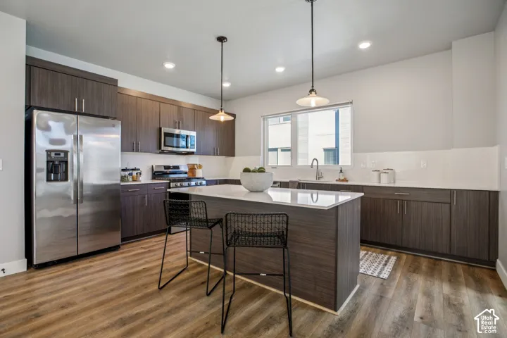 Kitchen featuring wood-finish flooring, dark wood-finish cabinetry, stainless steel appliances, a center island with a light-toned countertop, and recessed lighting