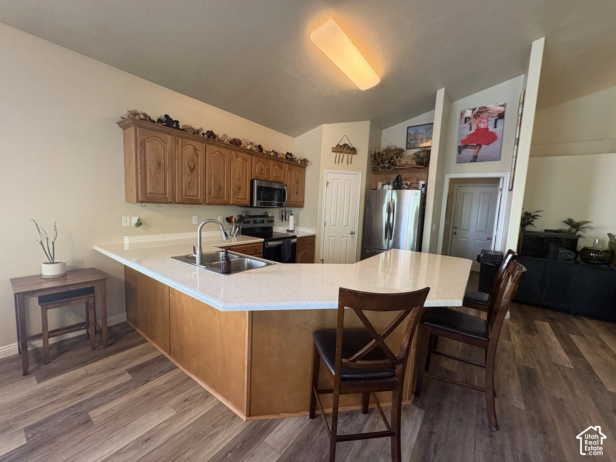 Kitchen featuring wood-finish flooring, an L-shaped island with a built-in sink, light-toned countertops, and wood cabinetry
