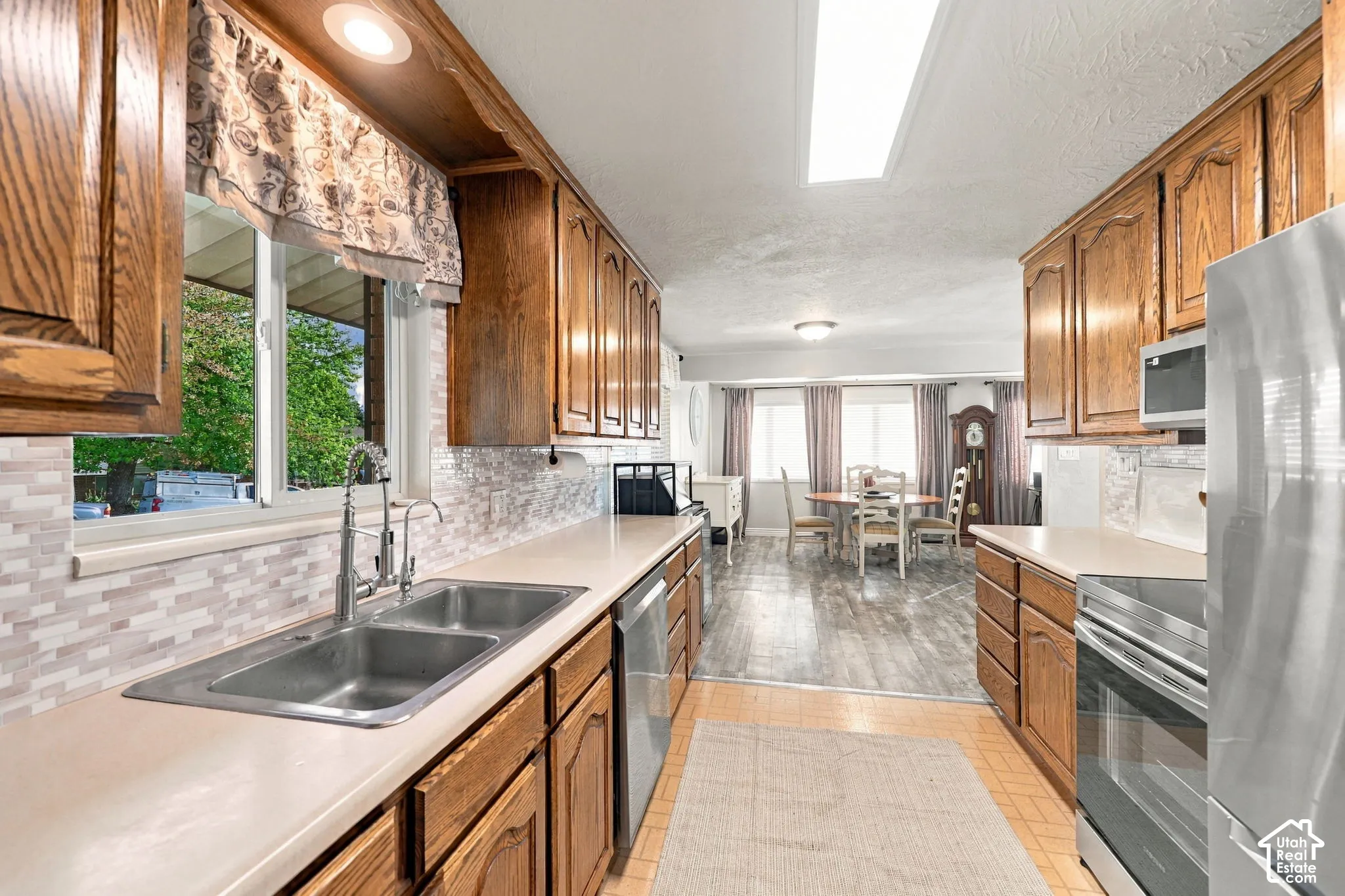 Galley kitchen with wood-finish cabinetry, solid surface countertops, a stainless steel double sink, and a tile backsplash